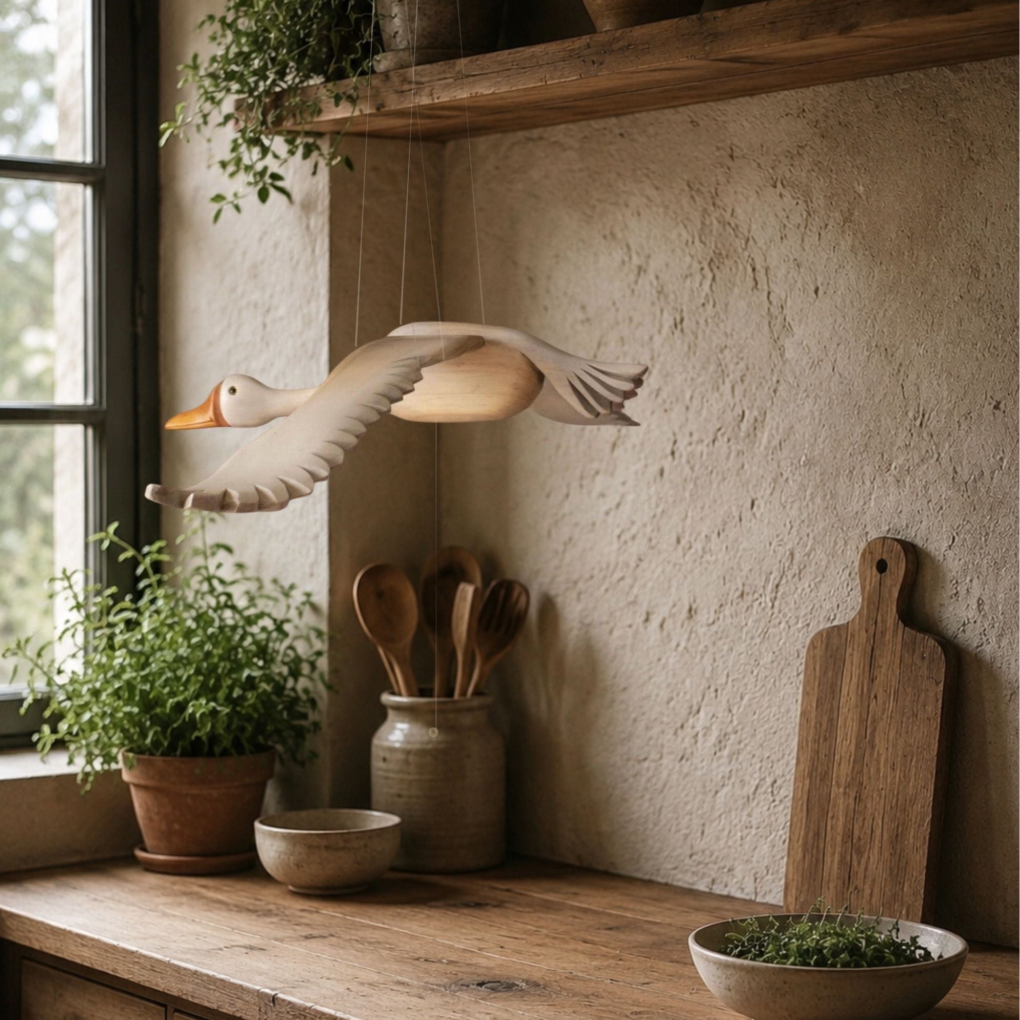 Decorative flying goose mobile hanging above a rustic kitchen counter with wooden cutting board, bowls, and plants.