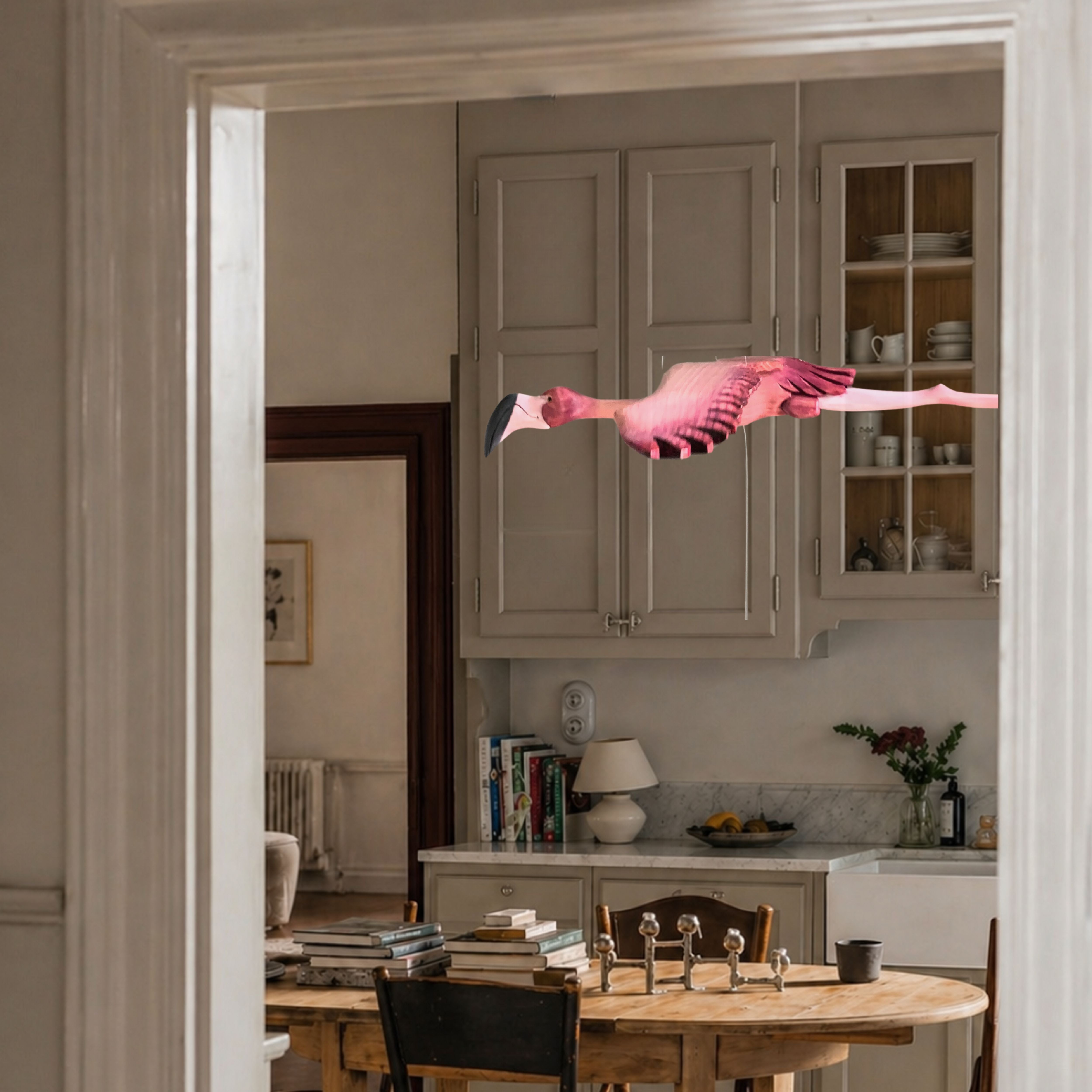 Hand-carved wooden flamingo mobile in flight through a bright country kitchen with wooden dining table and painted cabinets