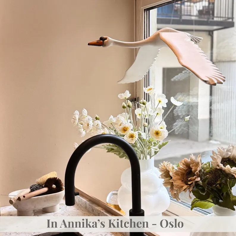 Hand-carved wooden swan mobile flying above a kitchen sink with flowers by the window